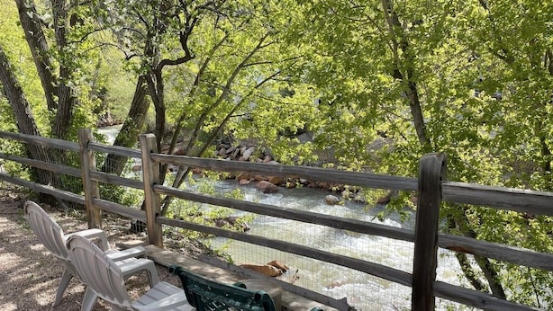 A wooden fence overlooking a river with rocks, surrounded by trees. There are two green benches and a white plastic chair. - Will it be ICE AXE, CRAWLER, NORDIC? 1200MGPS-Mtn Views! On River! BBQ & yard! (Ouray)
