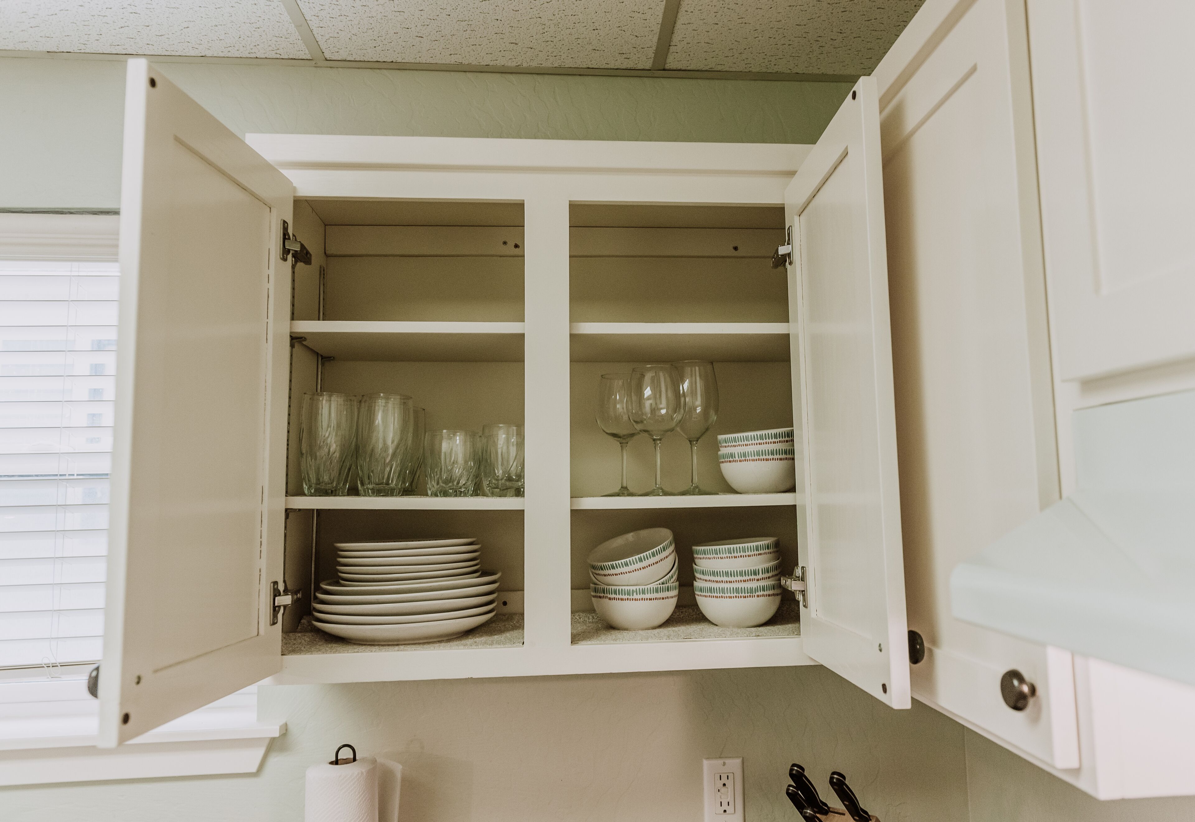 Una cocina con armarios blancos, un ventanal con persianas y una encimera con un juego de cuchillos.
