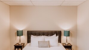 A neatly made bed with white linens, flanked by two bedside tables with lamps, in a room with a plain wall and a ceiling with a grid pattern. - Bay Breeze across from Waterfront (Eureka)
