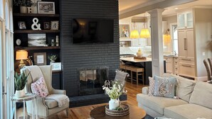 A modern living room with a fireplace, a ceiling fan, and a kitchen in the background. - SOUTHERN LIVING! Newly renovated! Private with Mountain views-sleeps 16 (Kingston)