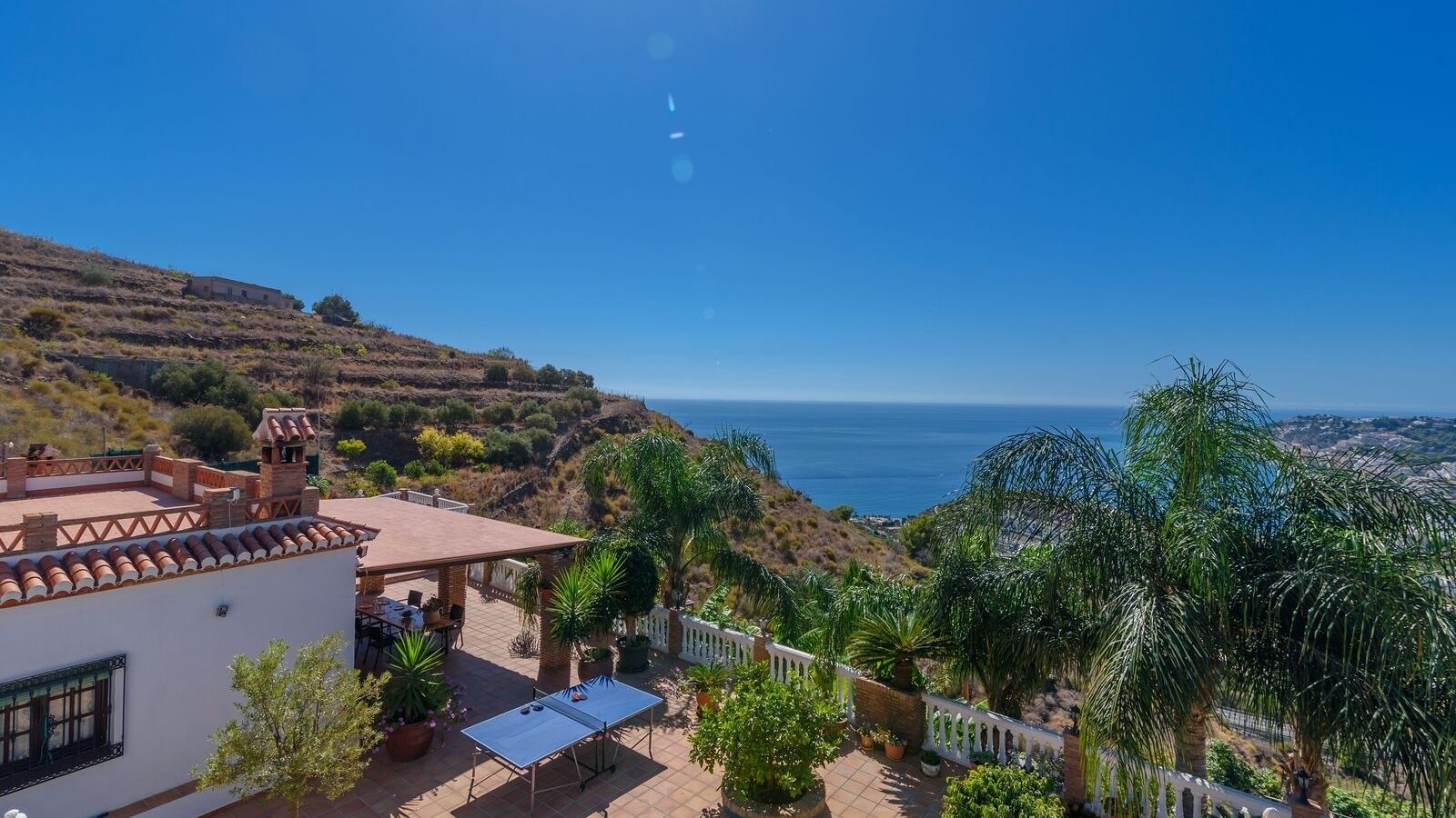 Une terrasse avec une table et des chaises, offrant une vue sur un paysage côtier avec des collines et la mer.