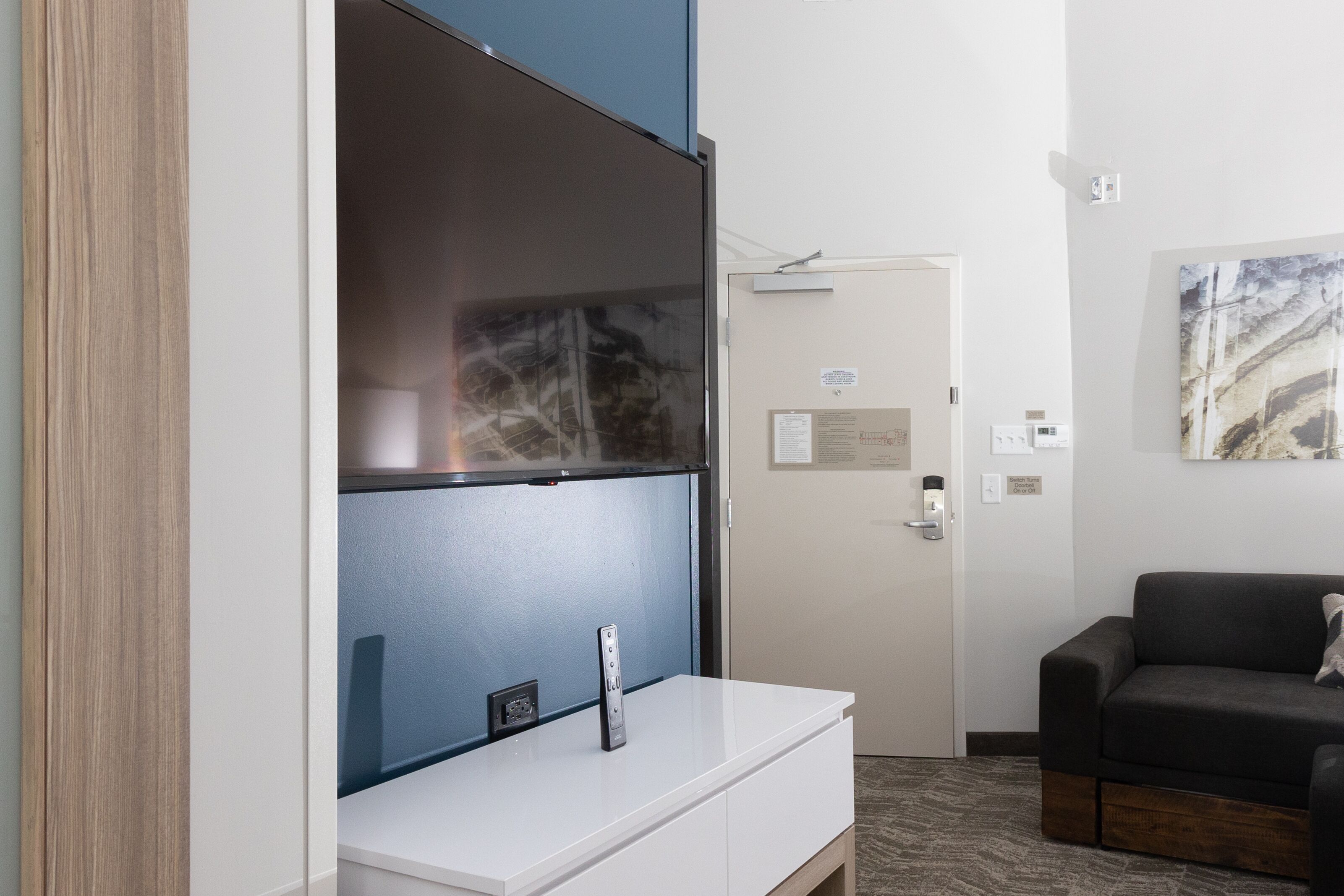 A modern hotel room with a large flat-screen TV mounted on a white cabinet, a dark-colored sofa, and a door with a small sign.