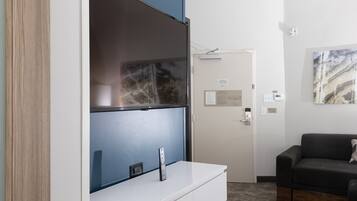 A modern hotel room with a large flat-screen TV mounted on a white cabinet, a dark-colored sofa, and a door with a small sign.