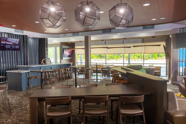 A modern restaurant with a central bar, multiple seating areas, and large windows offering a view of the outdoors.