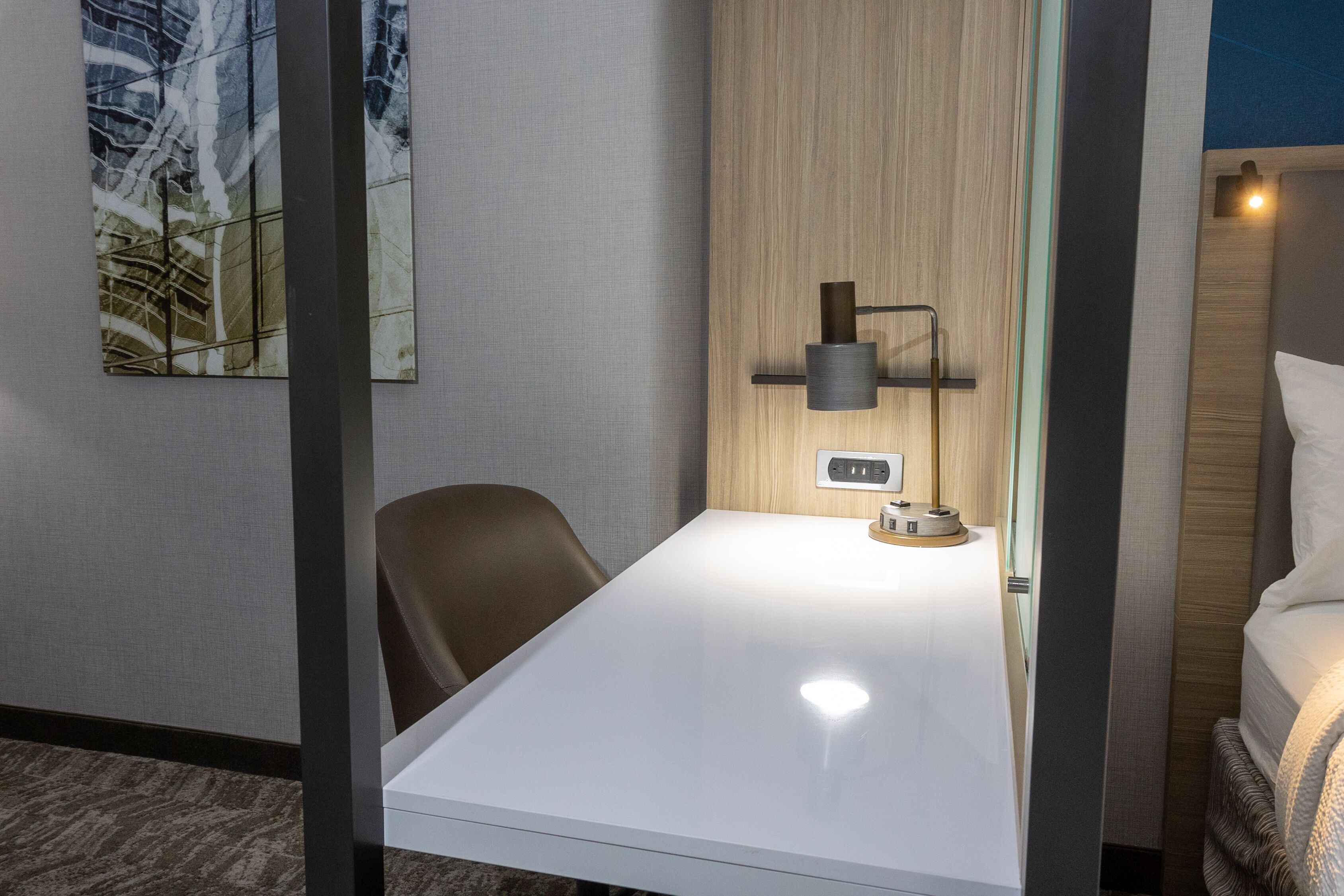Een hotelkamer met een wit bureau, een bruine stoel, een lamp en een bed met hoofdeinde.
