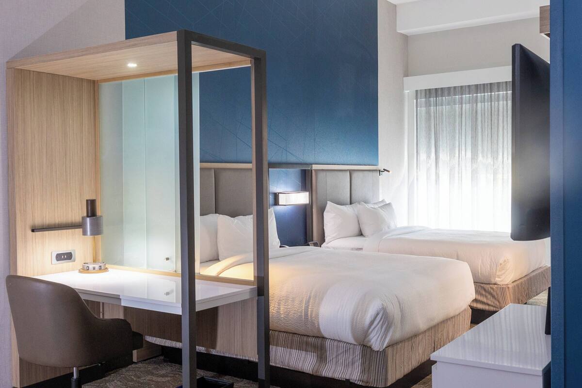A hotel room with a desk, two beds, a large mirror, and a TV.