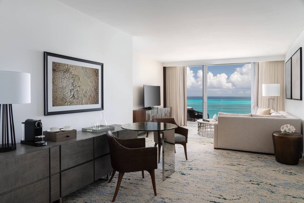 A modern hotel room with a dining area, a sofa, and a large window offering a view of the ocean.