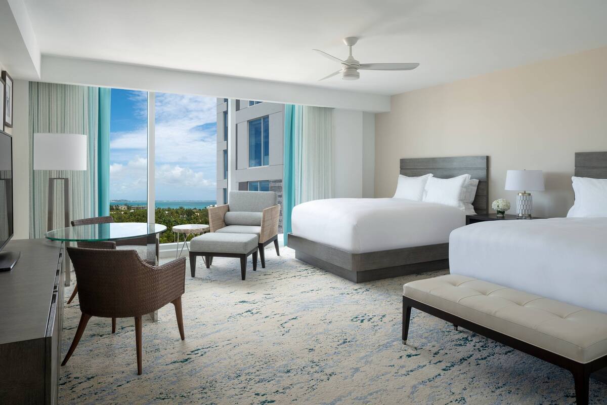 A hotel room with a large bed, a smaller bed, a desk with a chair, a round table with chairs, and a view of the ocean.