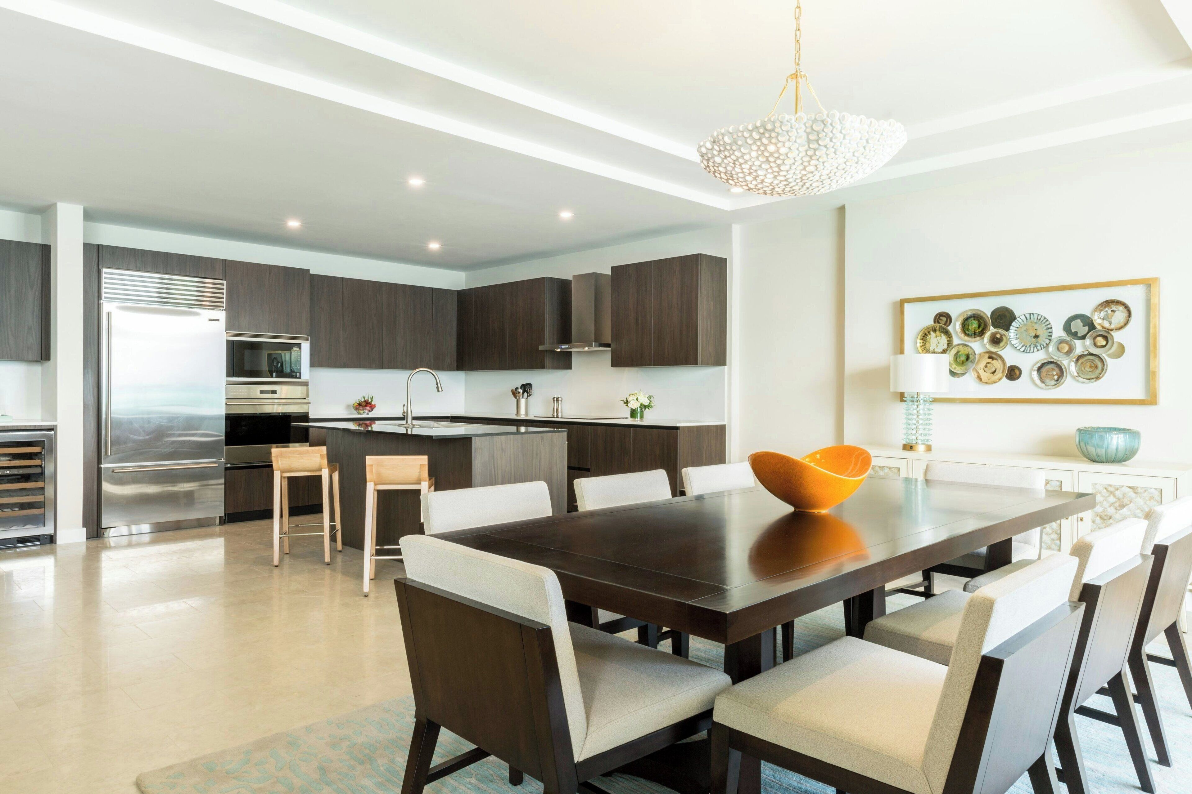 Una cocina moderna con armarios oscuros, un refrigerador de acero inoxidable y un comedor con mesa de madera oscura y sillas blancas.
