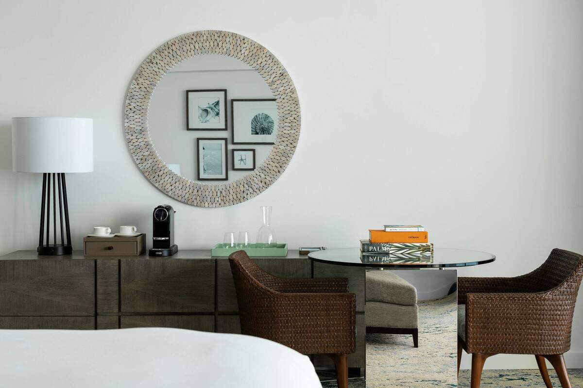 A modern hotel room with a round mirror, framed artwork, a coffee machine, a glass table, wicker chairs, and a bed.