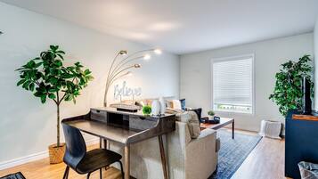 A modern living room with a dining area, a large potted plant, a wall-mounted light fixture, and a window with blinds.