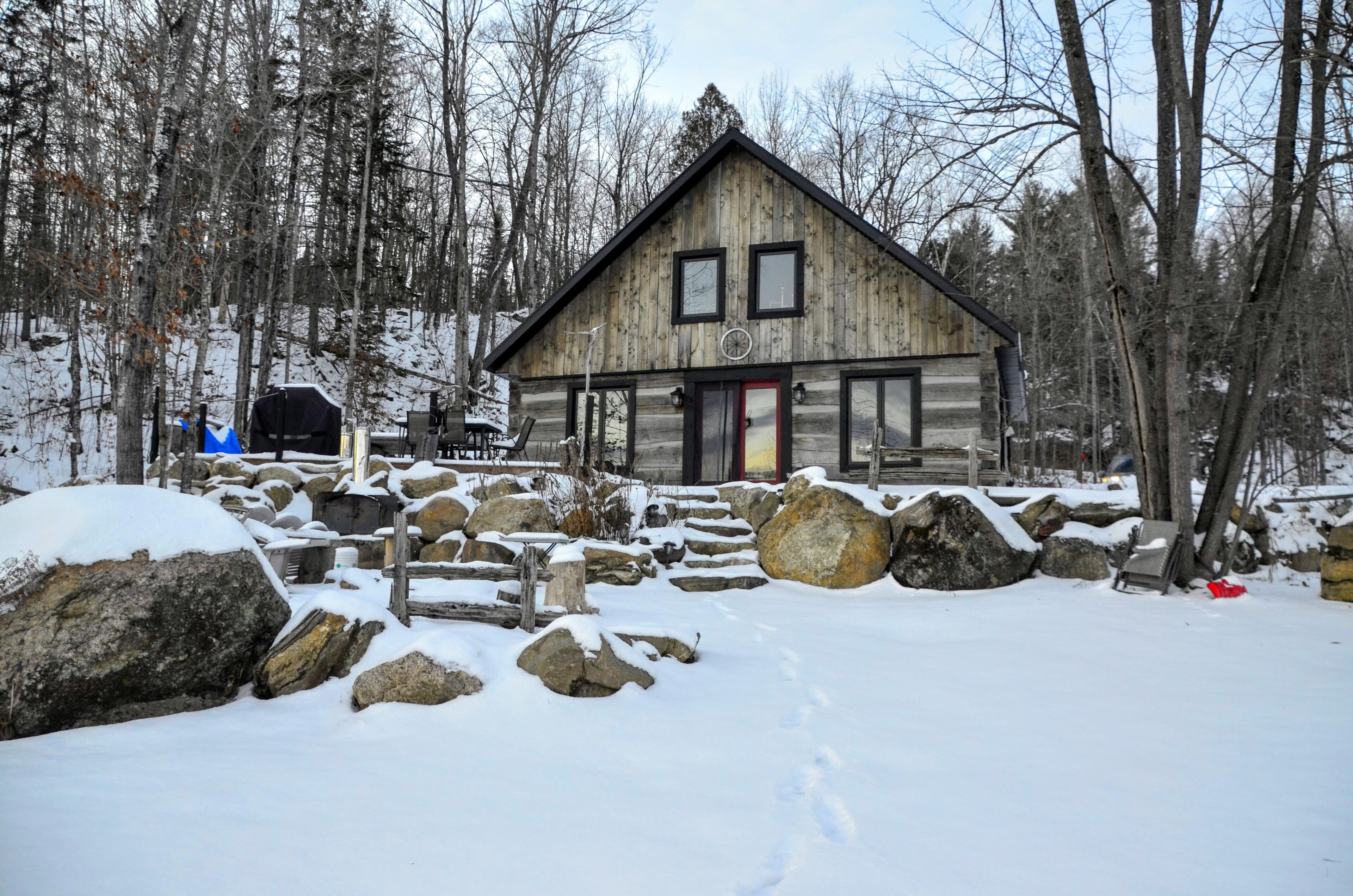 A wooden cabin with a red door, surrounded by snow-covered rocks and trees.