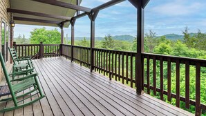 Un terrazzo in legno con ringhiera, poltrone a dondolo verdi e vista su un paesaggio boschivo.