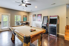 Une table de billard avec des billes, un ventilateur de plafond, des jeux d’arcade et une porte avec des stores.