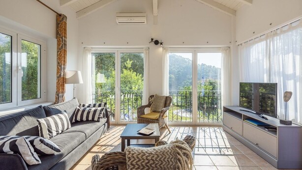 Un salon spacieux comprenant un canapé, des fauteuils, une table basse et un téléviseur posé sur un meuble. De grandes fenêtres offrent une vue sur un balcon et sur la verdure.