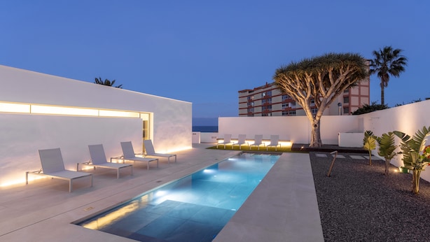 A modern outdoor pool area with lounge chairs and a tree, surrounded by a white building and a view of the ocean. - Remote workers paradise @ the Atlantic  (Las Caletillas)
