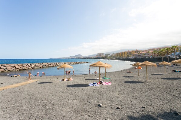 Ein Kiesstrand mit Sonnenschirmen, Personen, die sich sonnen, und einem Felssteg.