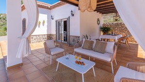 A sunlit patio with a white sofa, two wicker chairs, a white table with croissants, and a stone wall backdrop. - Villa Pitaya Frigiliana by Ruralidays (Málaga)