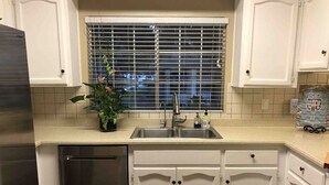 Una cocina con armarios blancos, lavavajillas de acero inoxidable, un lavamanos y una ventana con persianas.