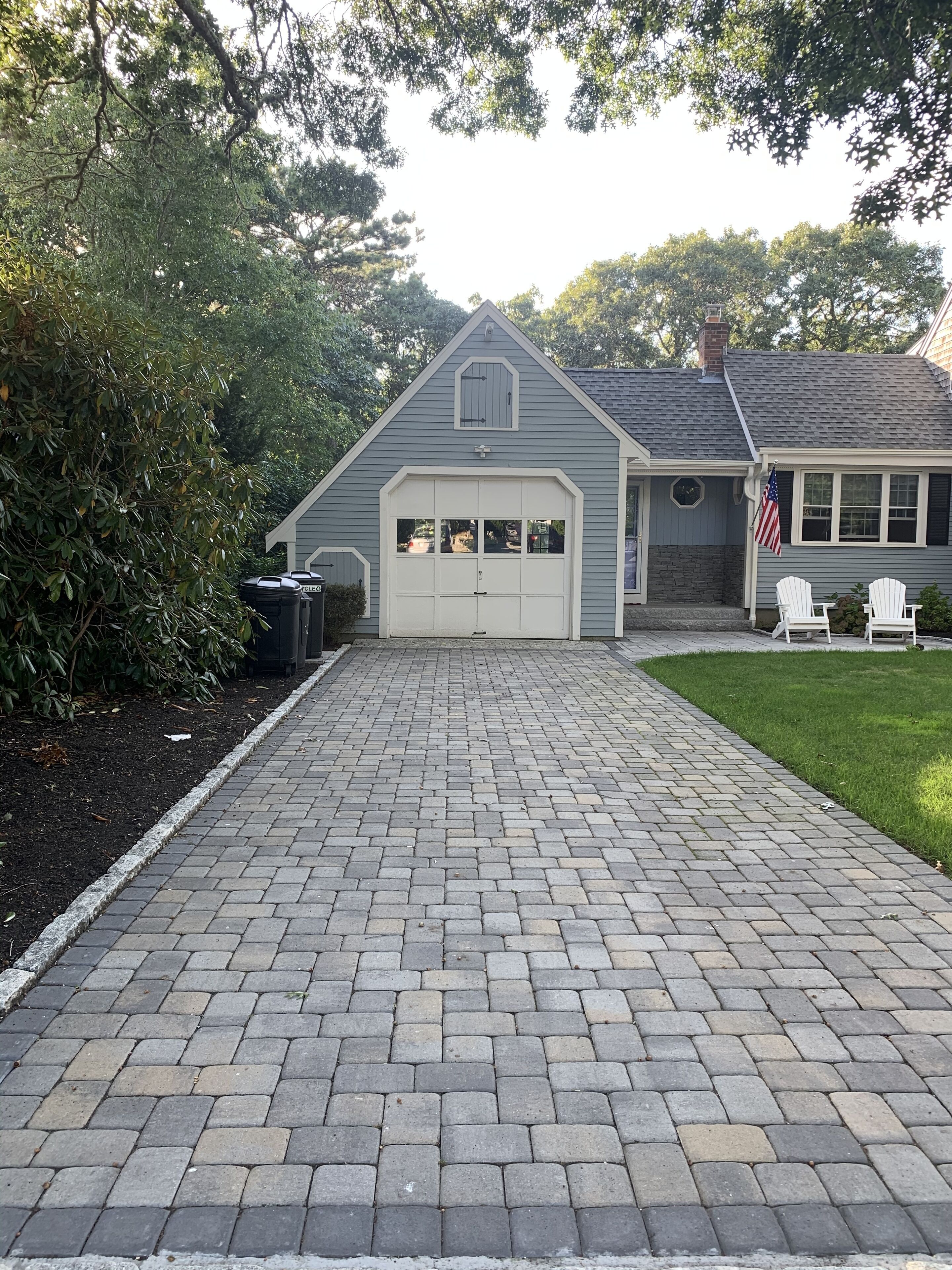 A house with a garage, a paved driveway, and a lawn with two white chairs.