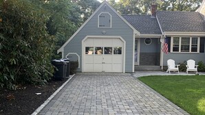 Una casa con garaje, un camino de entrada pavimentado y un césped con dos sillas blancas.