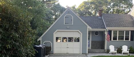 A house with a garage, a paved driveway, and a lawn with two white chairs.