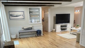 Un salón con suelo de madera, un banco, un televisor sobre un mueble bajo y una pared decorada con motivos náuticos.