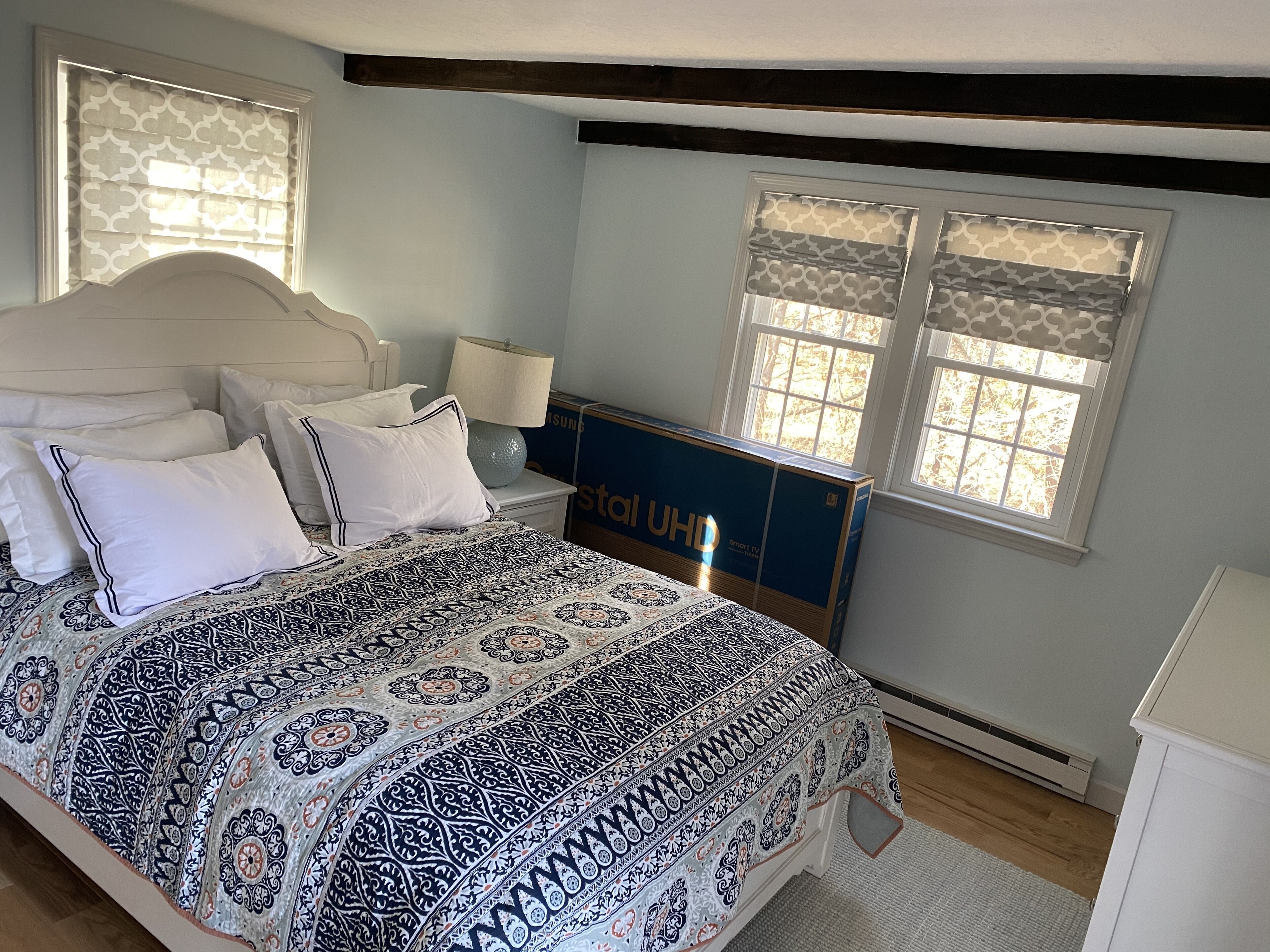 A bedroom with a large bed, a nightstand, a lamp, a TV stand, and two windows with patterned curtains.