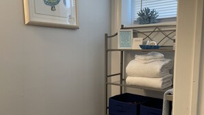 A bathroom with a toilet, a roll of toilet paper, a shelf with towels, and a framed picture of a turtle. - Bradley Beach Quaint Getaway (Bradley Beach)