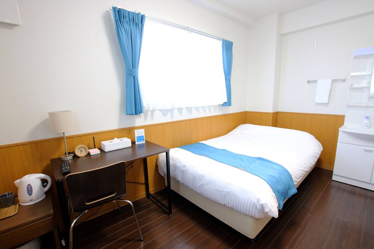 A hotel room with a bed, a desk with a lamp, a clock, and a window with curtains.