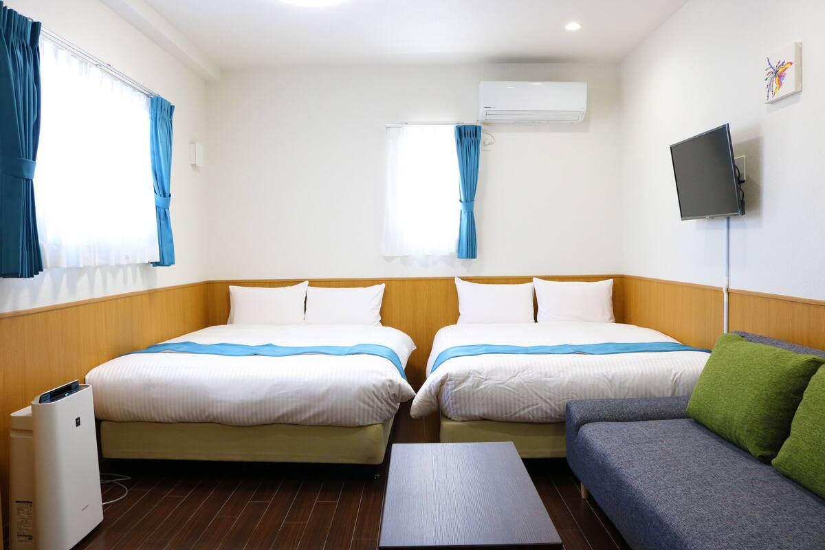 A hotel room with two beds, a sofa, a TV, and an air conditioner.