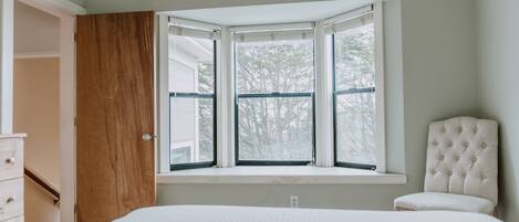 Quarto com uma cama grande, uma porta de madeira, uma janela com vista para árvores e uma cadeira.