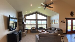 A spacious living room with a large flat-screen TV, a ceiling fan, and a view through large windows. - Beautiful Family Getaway with Breathtaking Teton Views (Tetonia)