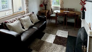 A living room with a black sofa, a dining table with chairs, a fireplace, and a large window with curtains.