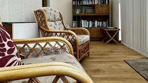 A room with a wooden floor, two wicker chairs with cushions, a bookshelf, and a small table.
