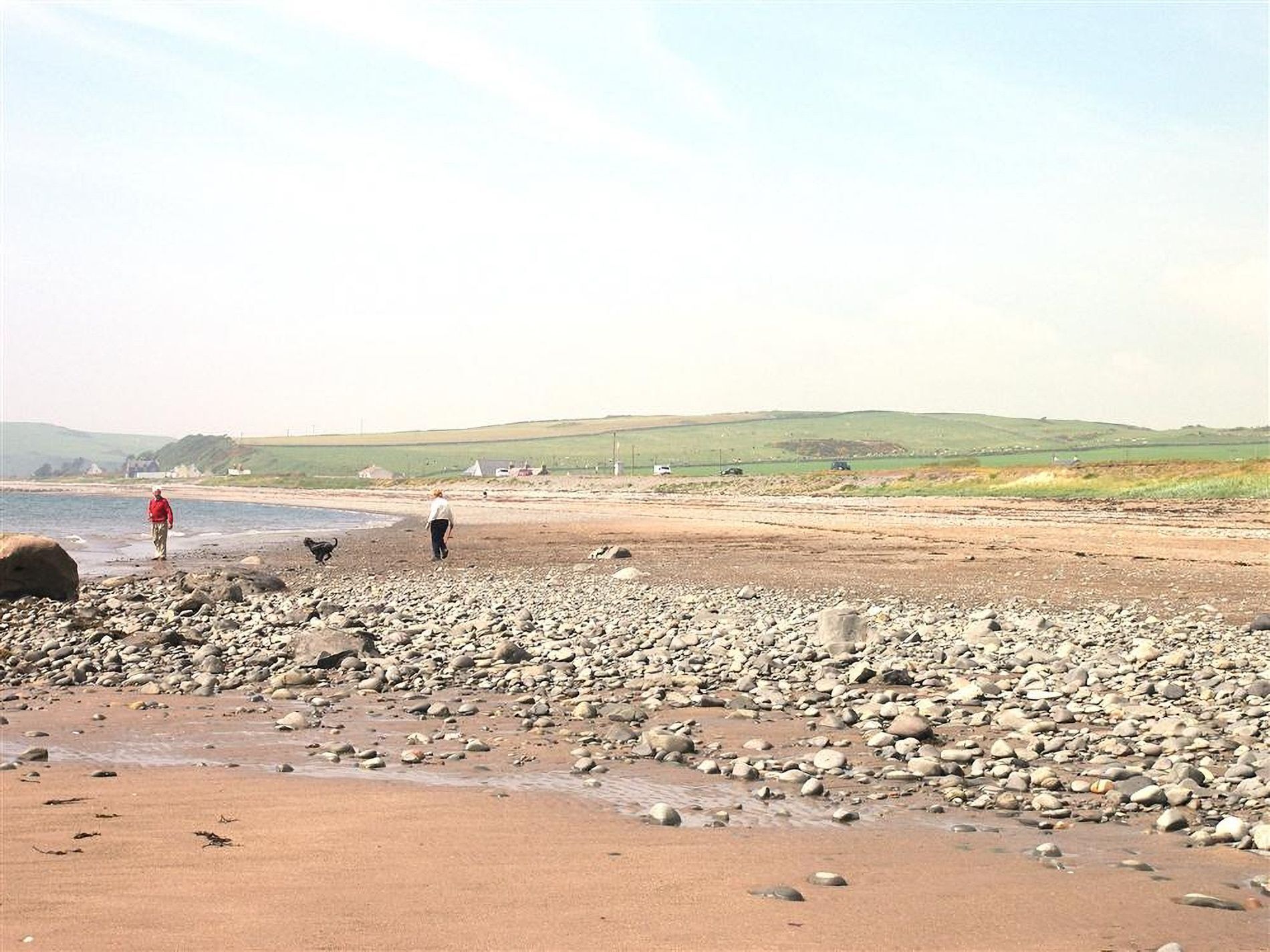 A pebbly beach with people walking and a dog, hills in the background, and a clear sky.