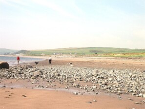 A pebbly beach with people walking and a dog, hills in the background, and a clear sky.
