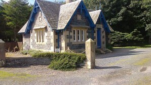 Une petite maison en pierre avec un toit à forte pente et des éléments décoratifs bleus, entourée de gravier et de verdure.