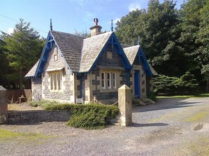 A stone cottage with a steeply pitched roof and blue trim, surrounded by gravel and greenery.