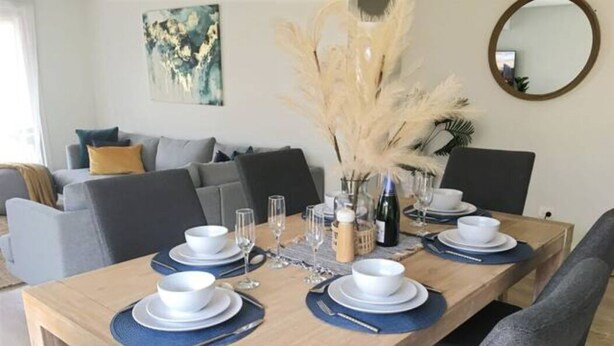 A dining table set with white plates, bowls, and cutlery, surrounded by grey upholstered chairs. - New Torquay Beaches Deakin Uni Hospitals (Armstrong Creek)