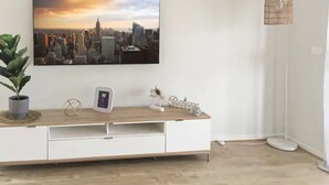 A modern living room with a cityscape painting, a white entertainment unit, a potted plant, and a floor lamp. - New Torquay Beaches Deakin Uni Hospitals (Armstrong Creek)