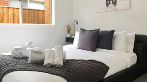 A neatly made bed with white and dark grey bedding, a black headboard, and a small bedside table with a lamp. - New Torquay Beaches Deakin Uni Hospitals (Armstrong Creek)