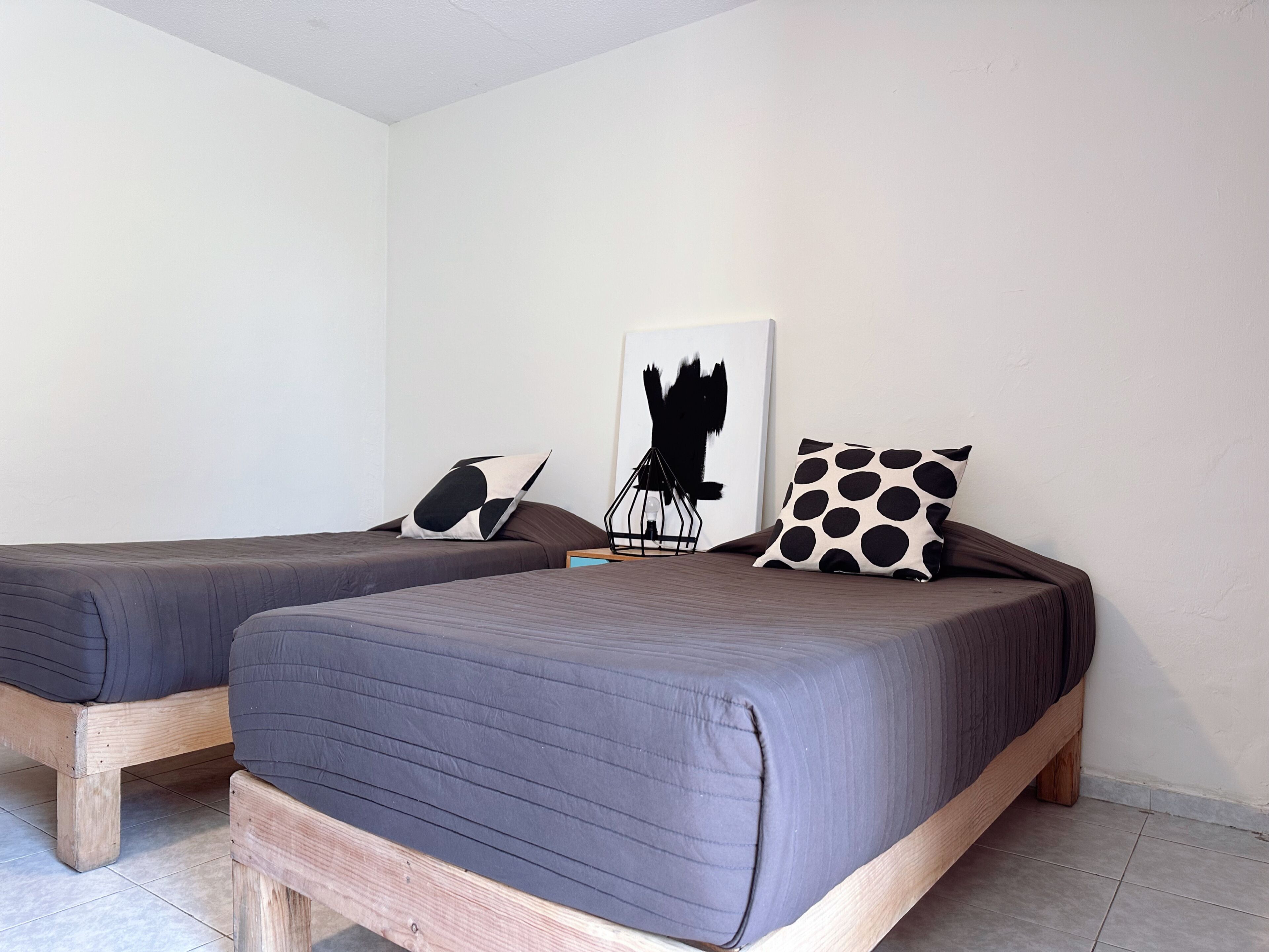 A room with two single beds, each with a wooden frame and dark bedding. There is a black and white abstract painting on the wall and a black and white polka dot pillow on one bed.