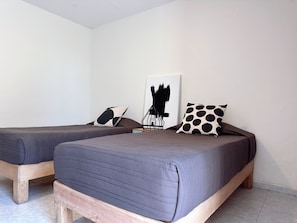 Una habitación con dos camas individuales, cada una con estructura de madera y ropa de cama oscura. Hay un cuadro abstracto en blanco y negro en la pared y un cojín blanco y negro con lunares en una de las camas.