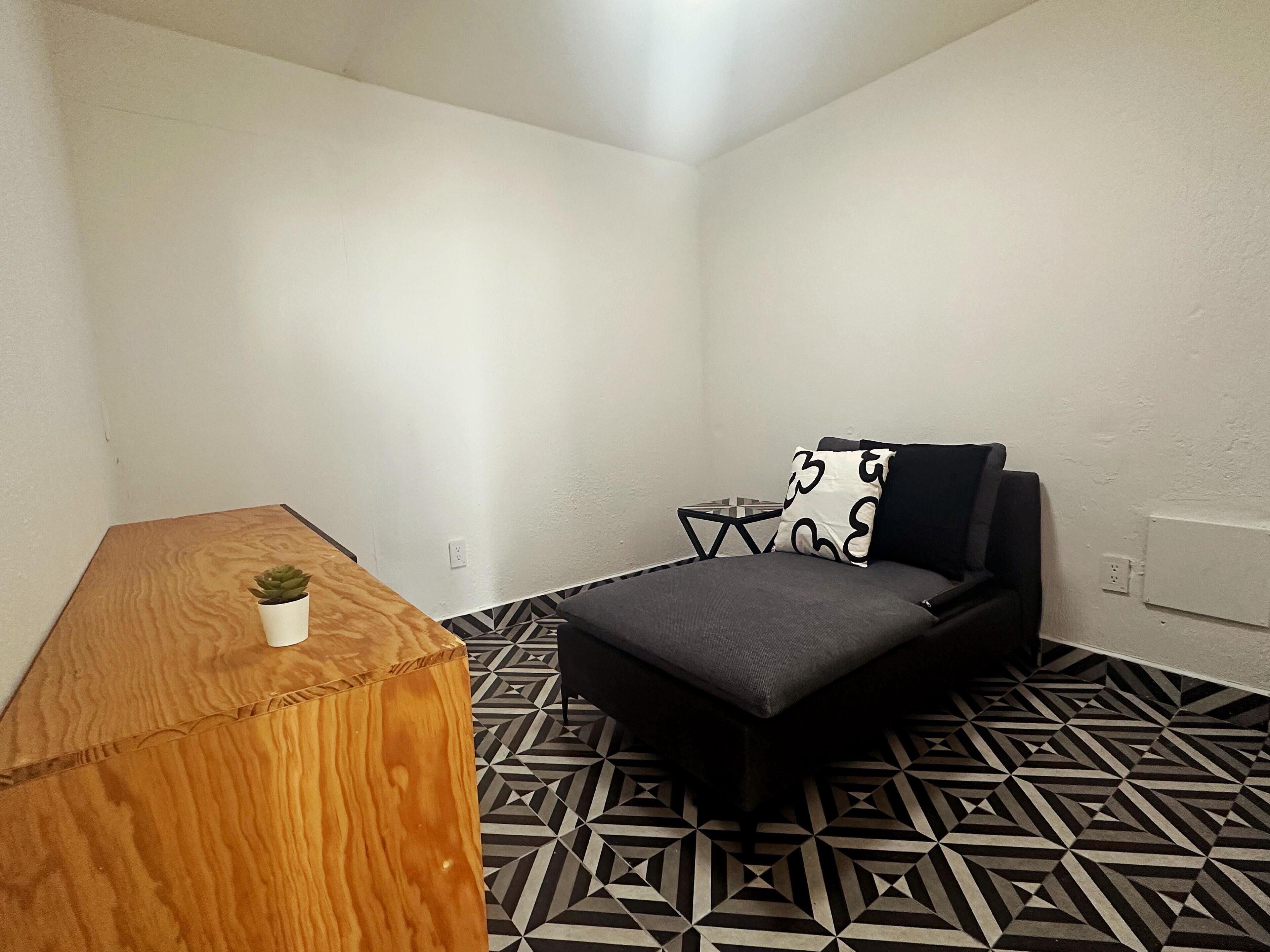 Una habitación pequeña y moderna con una mesita de madera, una planta pequeña en maceta, un sofá con estampado blanco y negro y un suelo con diseño geométrico.