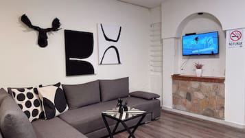 Una sala de estar moderna con un sofá gris, una mesa de centro y arte abstracto en la pared. Un televisor está montado en la pared, y se puede ver un cartel de prohibido fumar.
