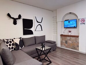 Una sala de estar moderna con un sofá gris, una mesa de centro y arte abstracto en la pared. Un televisor está montado en la pared, y se puede ver un cartel de prohibido fumar.