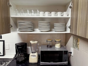 Una cocina con armarios blancos, un microondas, una licuadora y estantes con platos y tazas apilados.