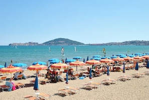 A beach with rows of sun loungers and umbrellas, a clear sky, and sailboats on the water.