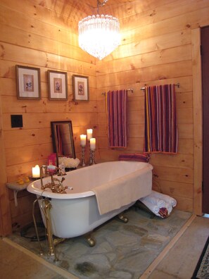 A wooden cabin bathroom with a clawfoot bathtub, framed pictures on the wall, and a chandelier.